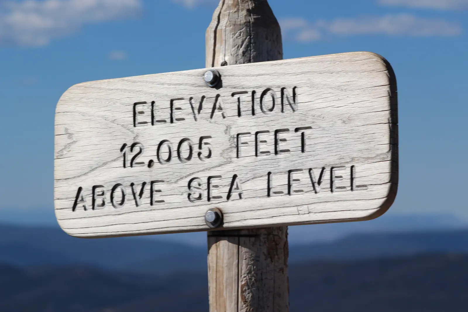 A weathered wooden sign reading 'Elevation 12,005 feet above sea level' against blue sky and distant blue ridges