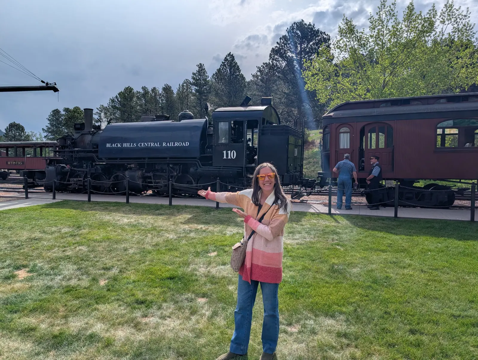 A visitor in a striped cardigan gestures toward a black Black Hills Central Railroad steam locomotive numbered 110 and a red passenger car on a grassy platform under ponderosa pines