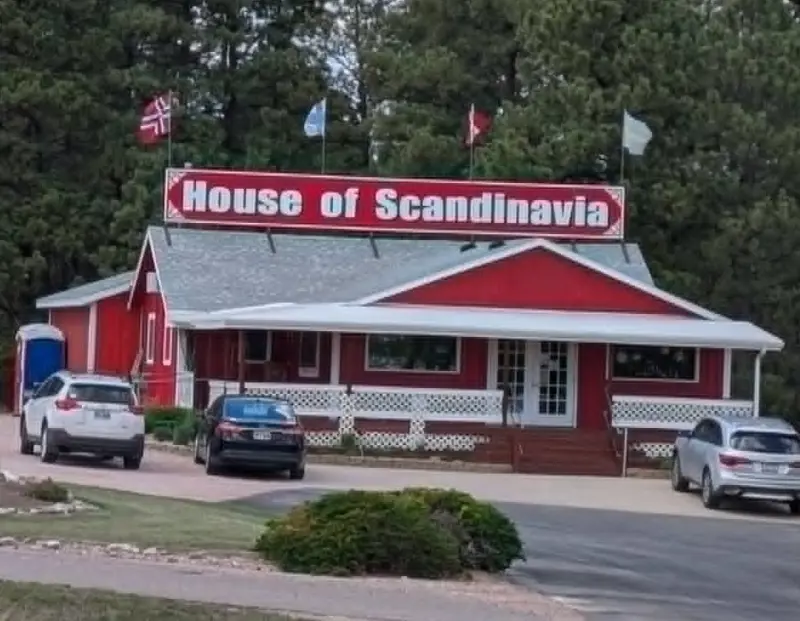 A red barn-style shop with white trim and a large 'House of Scandinavia' sign, Nordic flags flying from the roof, with cars parked in front under pine trees
