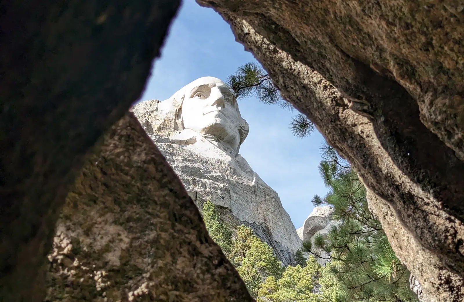 George Washington's carved granite face seen through a narrow gap between two boulders, framed by pine branches and blue sky