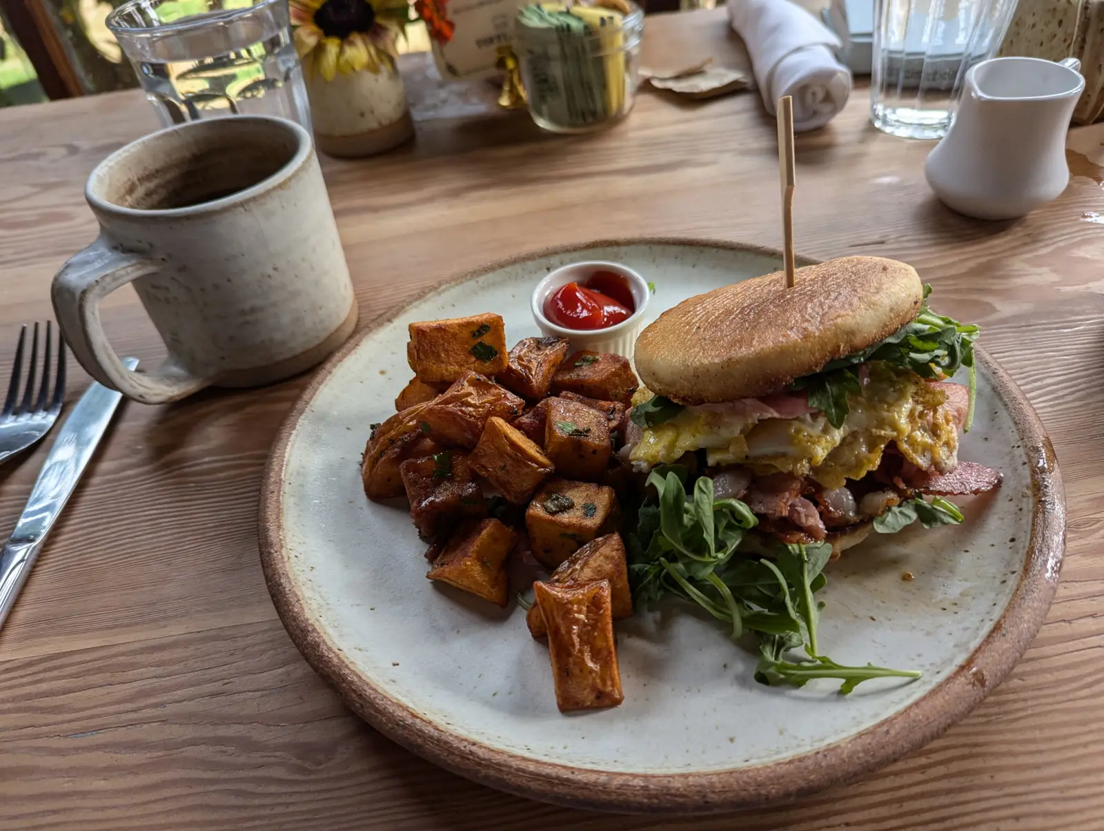 A breakfast sandwich stacked with bacon and arugula beside roasted sweet potatoes on a ceramic plate, with a mug of coffee on the wooden table