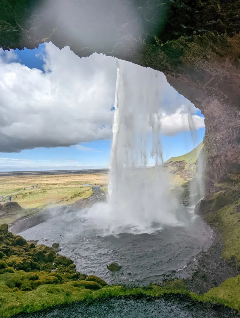 View from the mossy cave behind Seljalandsfoss, looking out through the falling curtain of water at the green plain and walking path beyond