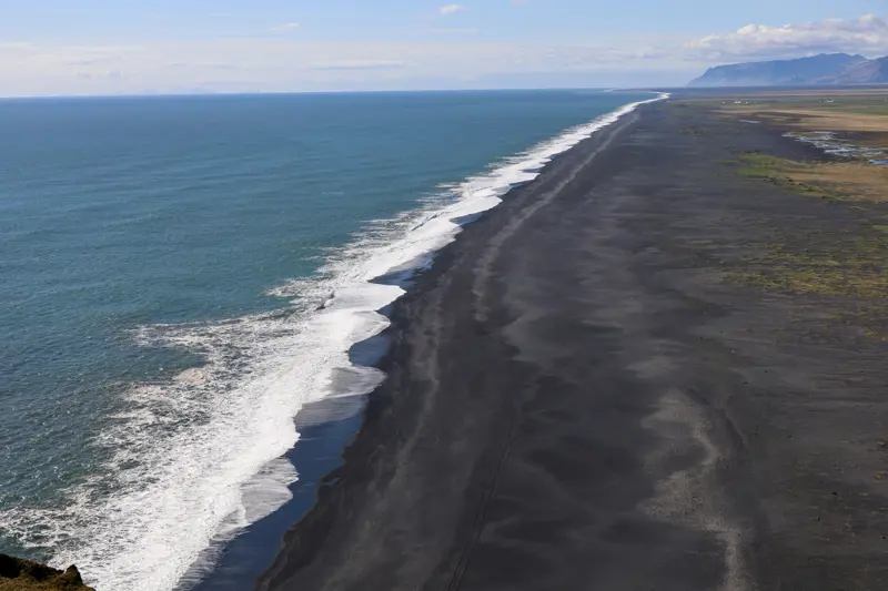 Aerial view along a long, straight black-sand beach with white surf on the left and flat green farmland on the right, mountains and islands on the horizon