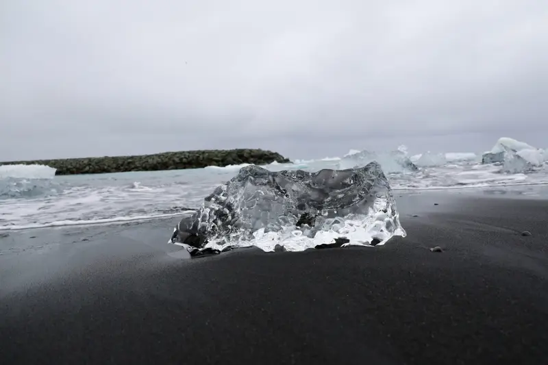 Clear, sculpted chunk of glacial ice sitting on wet black sand with waves and more ice fragments washing in behind it