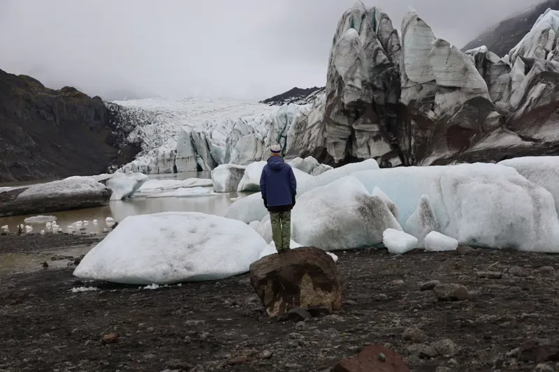Person in a blue jacket standing on a boulder facing a glacier tongue of ash-streaked ice, with icebergs grounded in a muddy meltwater lagoon