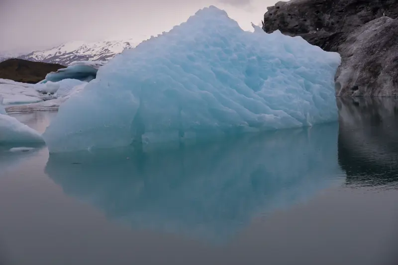 Large translucent blue iceberg with a rippled surface floating in still gray water, reflected below, with snow-capped mountains behind