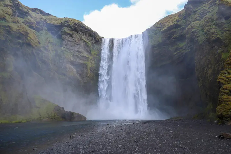 Skógafoss thundering off a wide green cliff into a rocky riverbed, with mist filling the amphitheatre of moss-covered rock