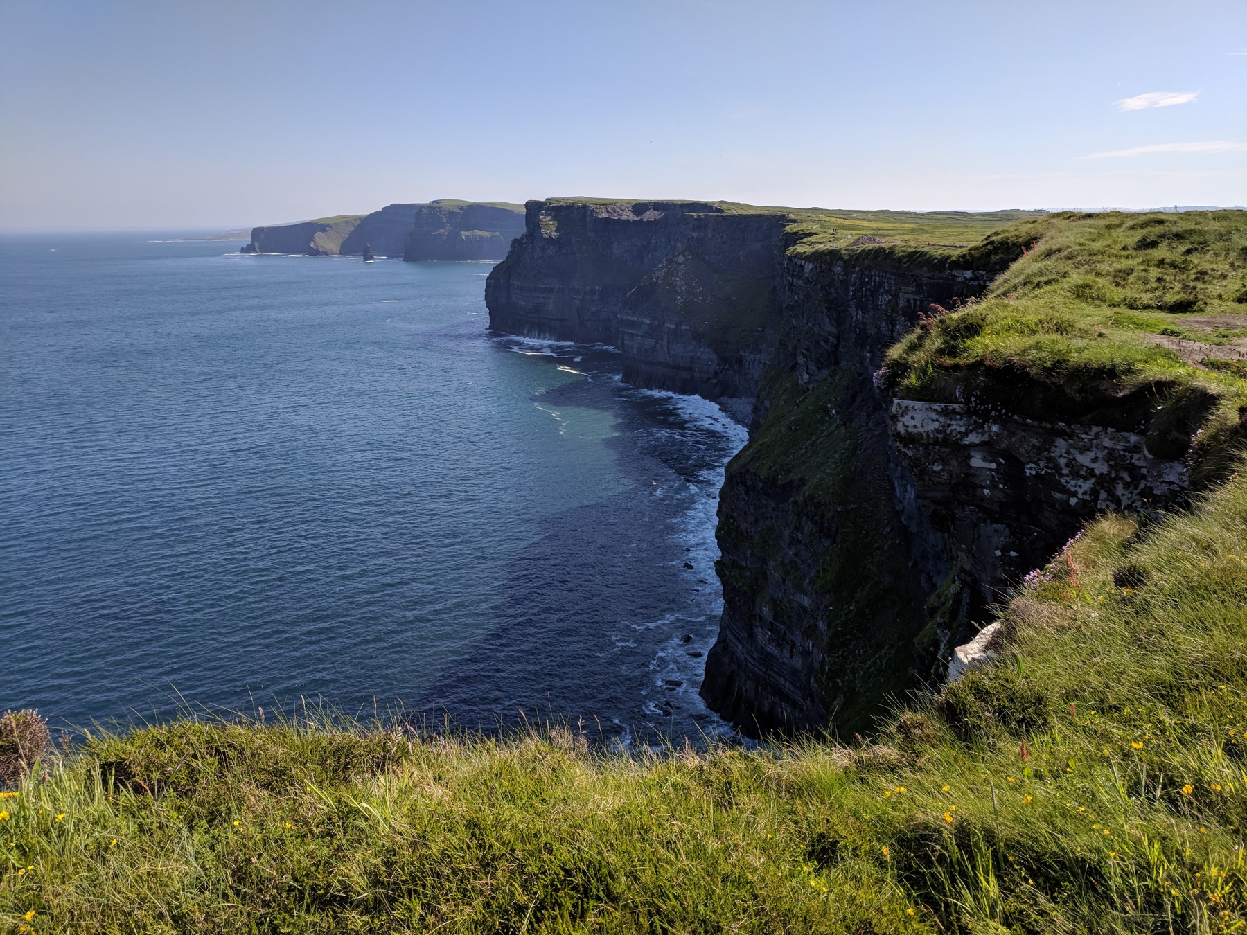 Sheer dark cliffs dropping into deep blue Atlantic water with a lone sea stack and a distant headland under a clear sky