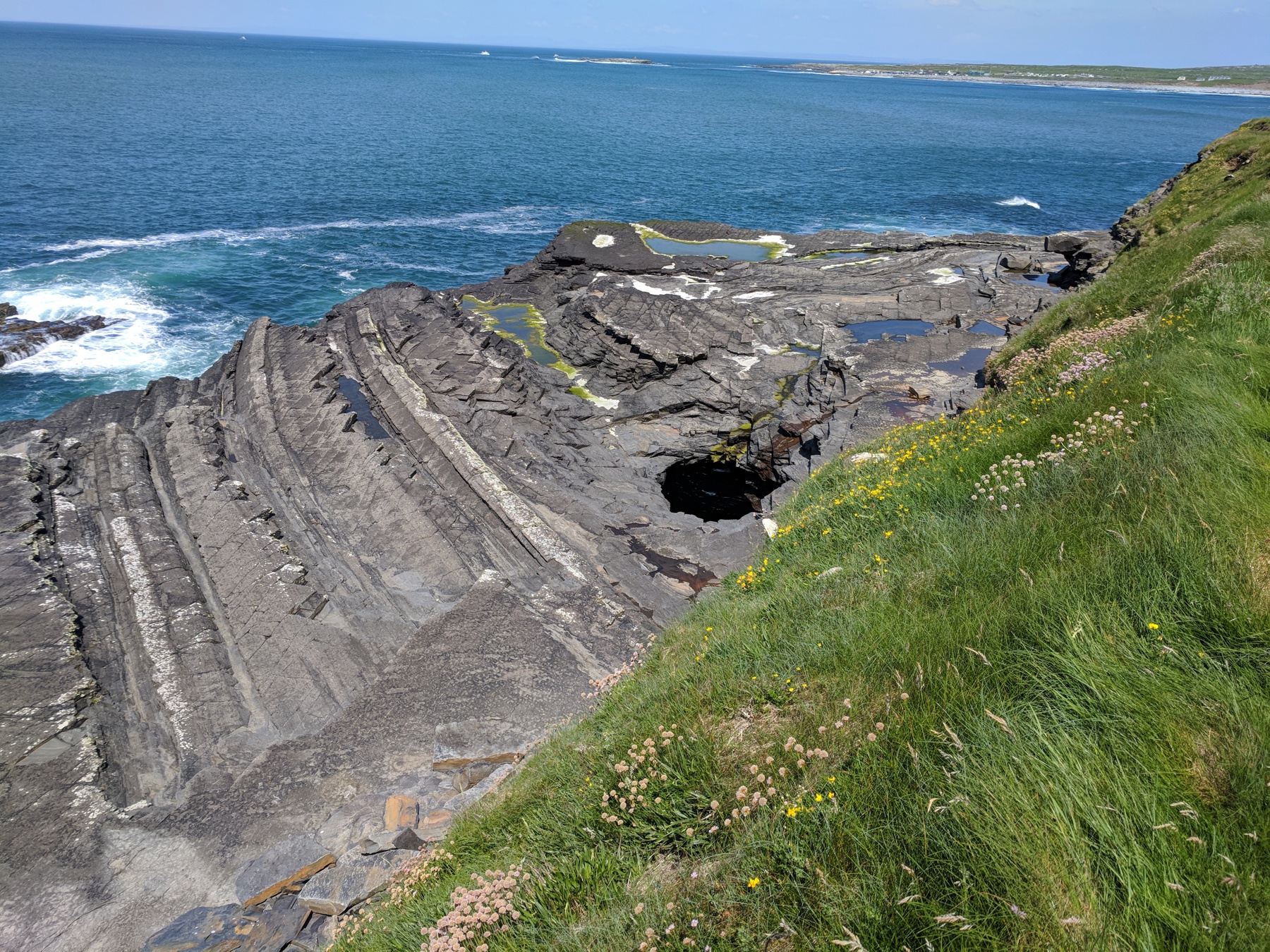 Tilted grey flagstone ledges with tide pools sloping into churning turquoise surf, wildflowers on the grassy bank above and a distant village across the bay