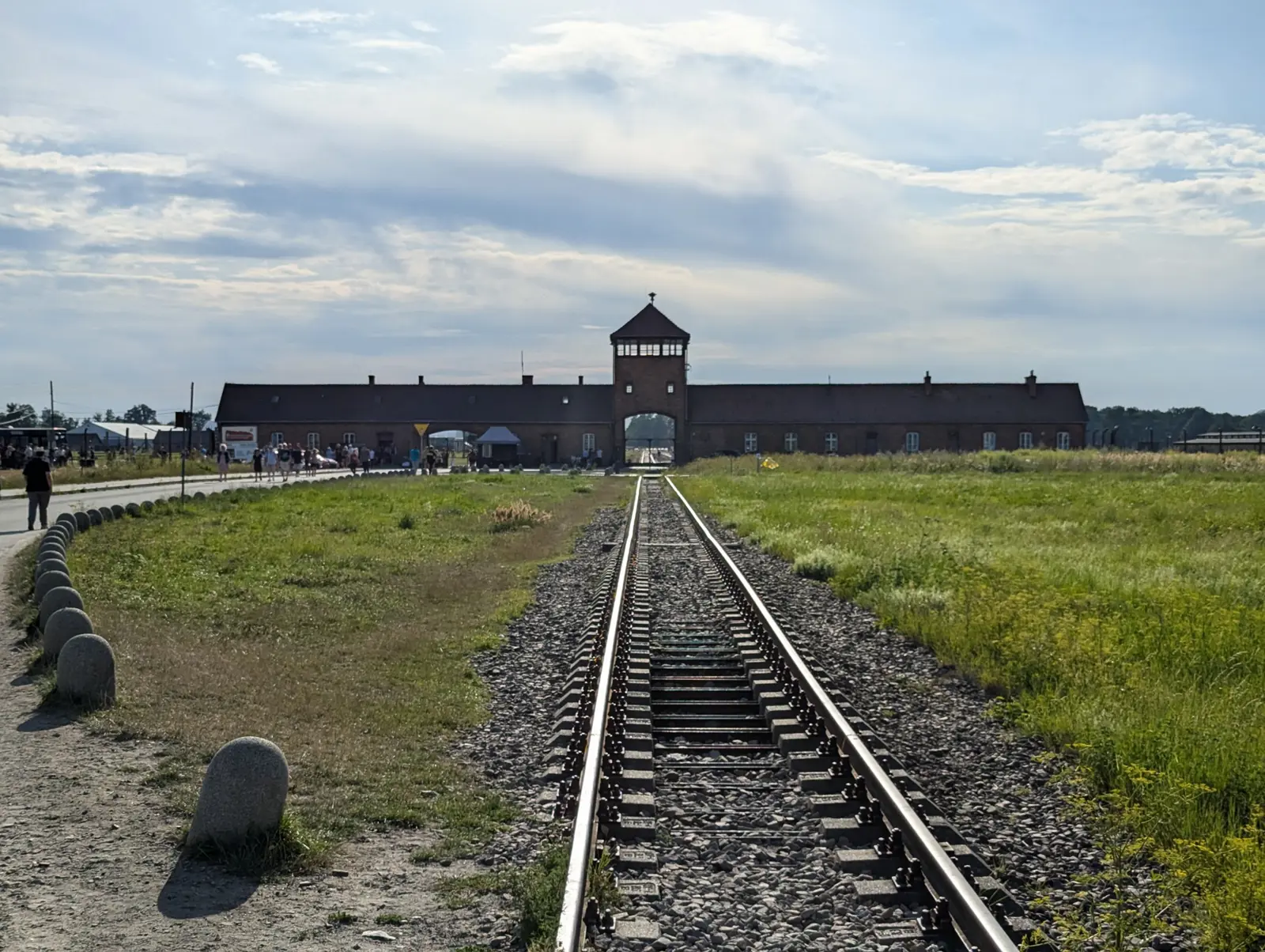 The rail track running straight through the grass toward the brick gatehouse of Birkenau under a wide, pale sky