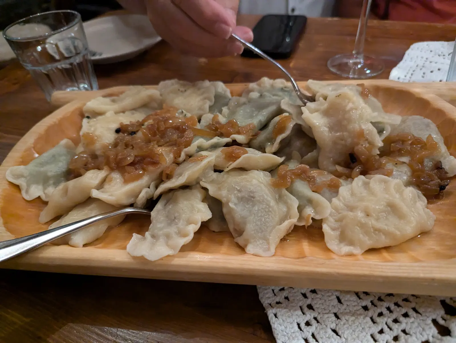 A wooden platter heaped with boiled pierogi topped with caramelized fried onions, forks digging in