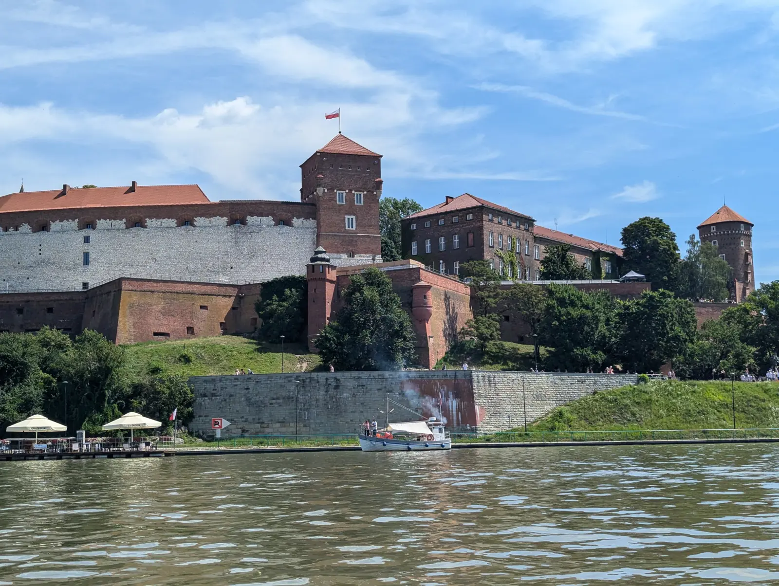 Wawel Castle's towers and red-brick ramparts seen from water level on the Vistula, with café umbrellas and a small boat along the bank