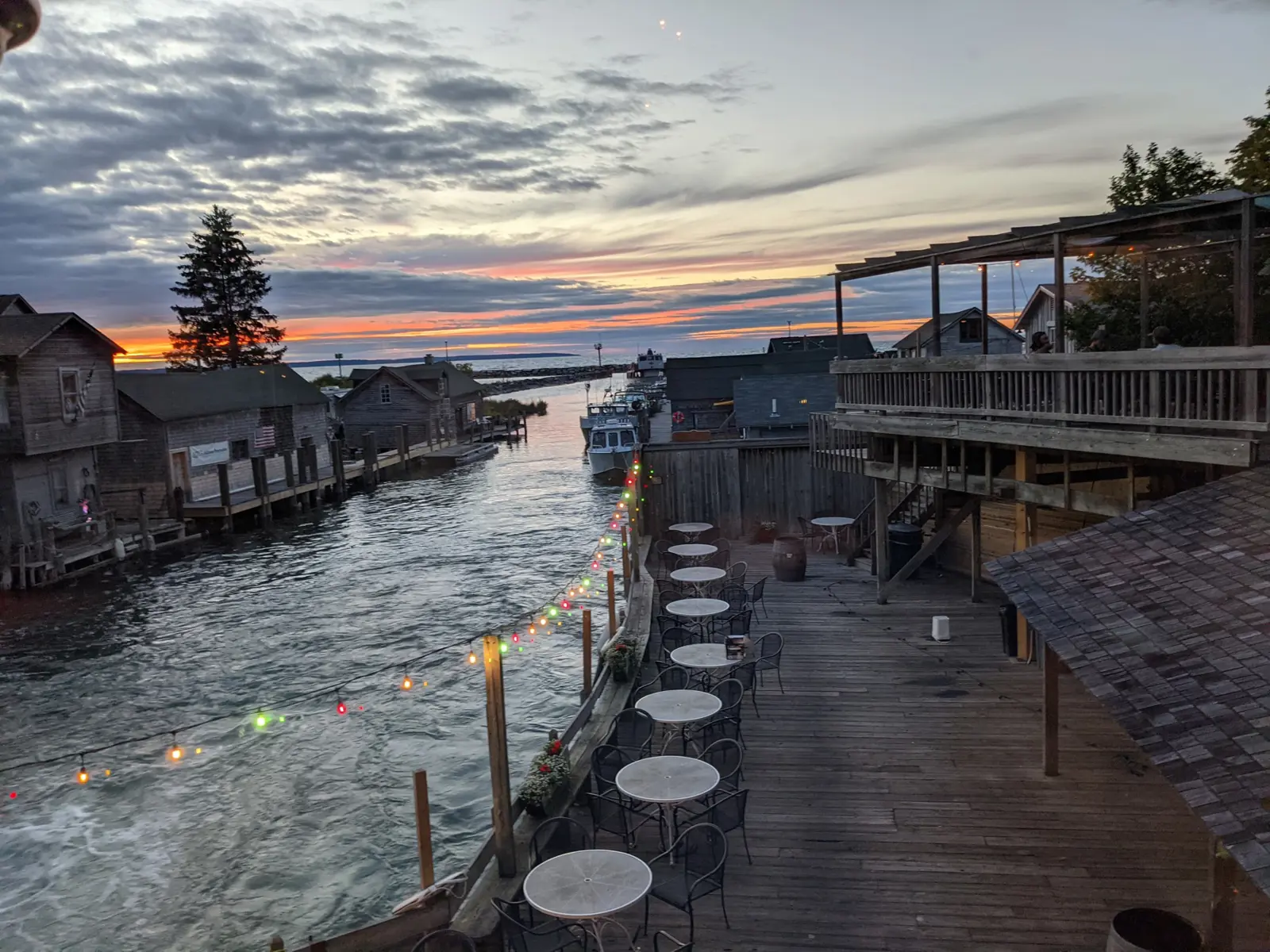 String lights and café tables on a wooden deck above a river channel at sunset, with weathered fishing shanties and a docked tug across the water
