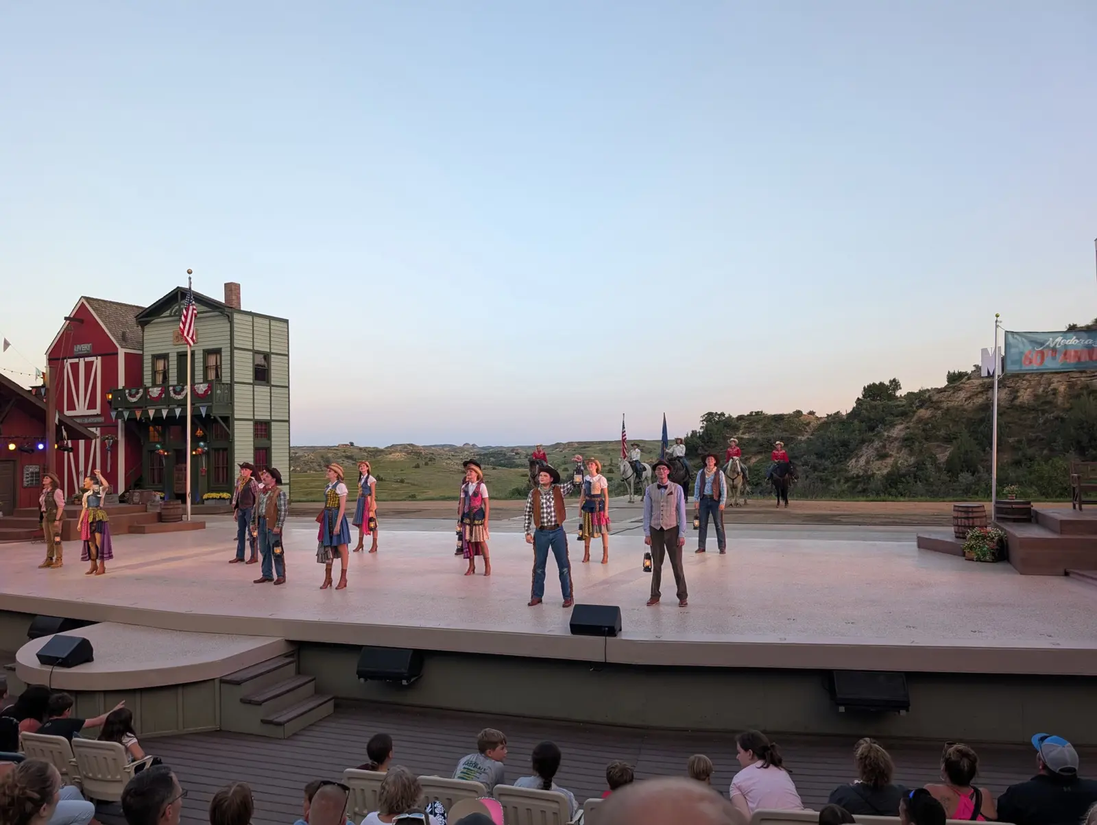 Performers in Western costume lined up on the Burning Hills Amphitheatre stage at dusk, with a red barn set piece and the badlands behind them