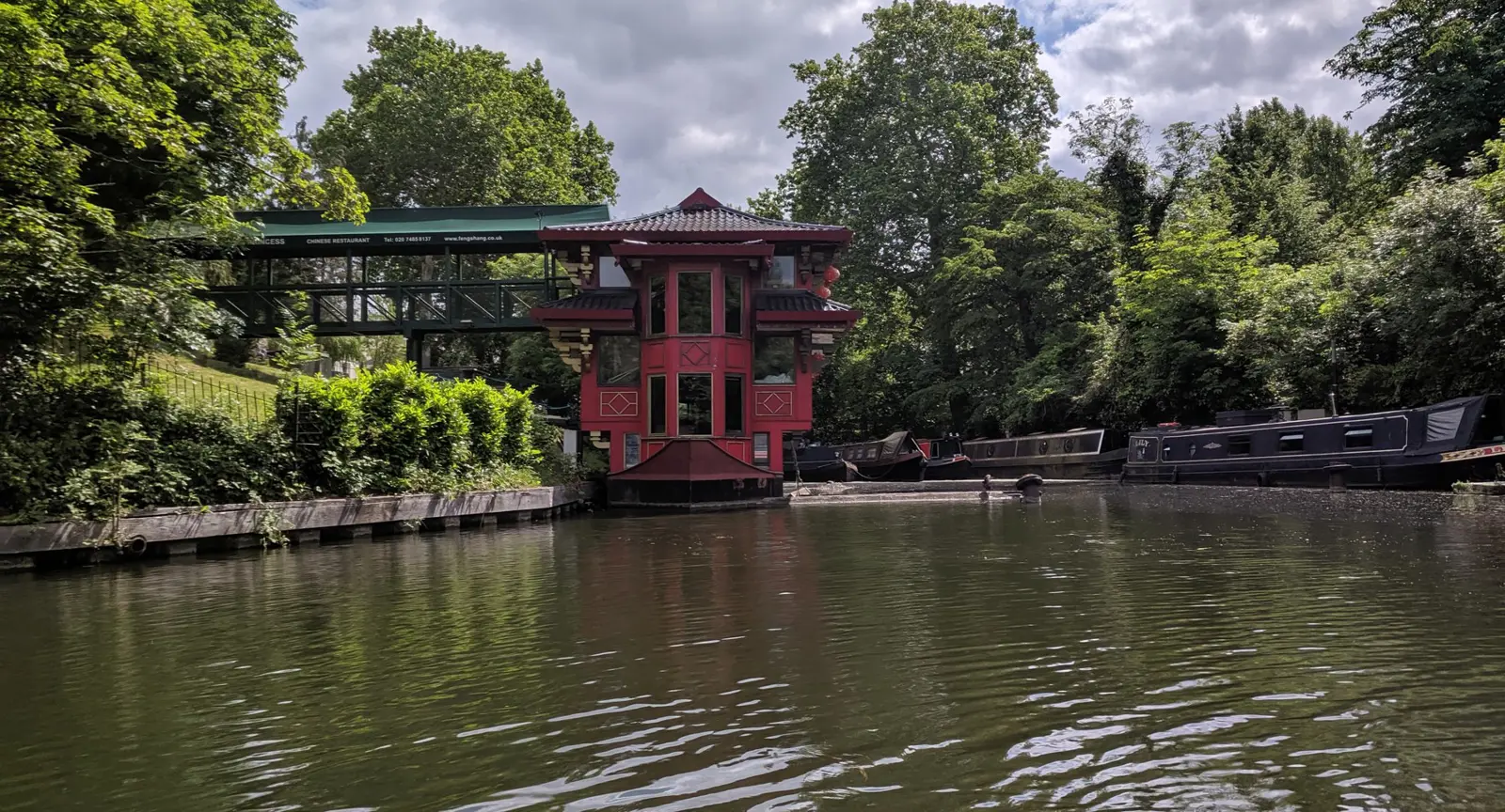 A red three-story pagoda-style floating restaurant moored on a tree-lined canal basin with black narrowboats alongside