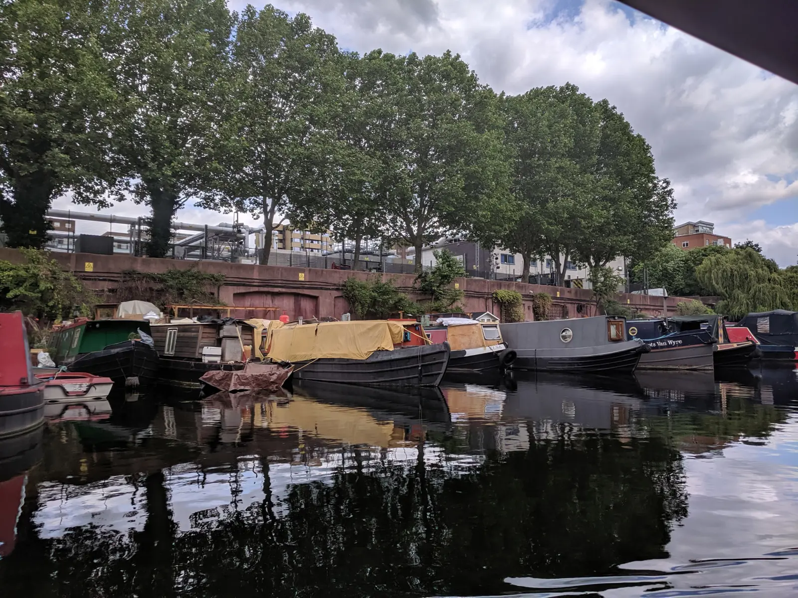 A row of weathered liveaboard narrowboats moored under plane trees on the Regent's Canal, reflected in still water