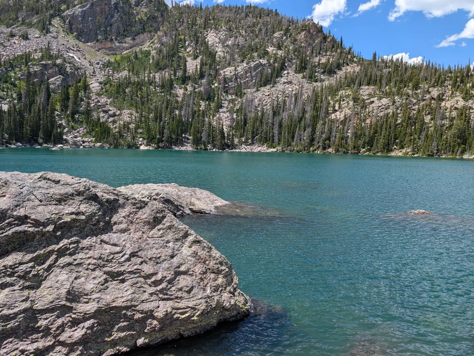 The milky-turquoise water of Lake Haiyaha behind a large granite boulder, below a forested rocky slope