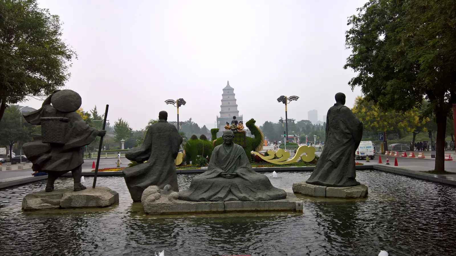 Stone statues of robed Tang-era figures standing in a fountain pool, with the tiered Giant Wild Goose Pagoda rising in the haze behind