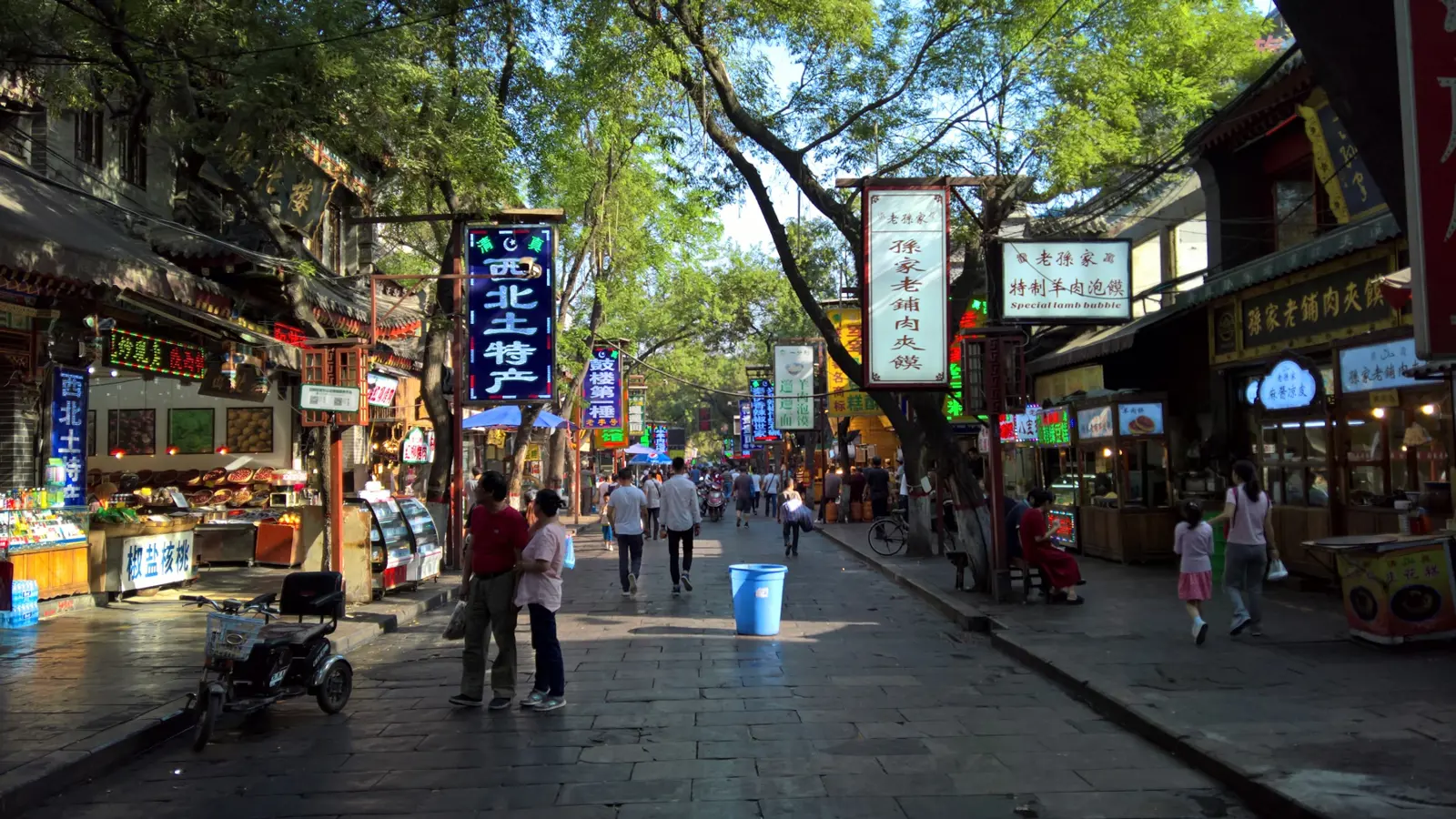 A tree-shaded Muslim Quarter street lined with vertical shop signs and food storefronts, with people strolling in the morning light