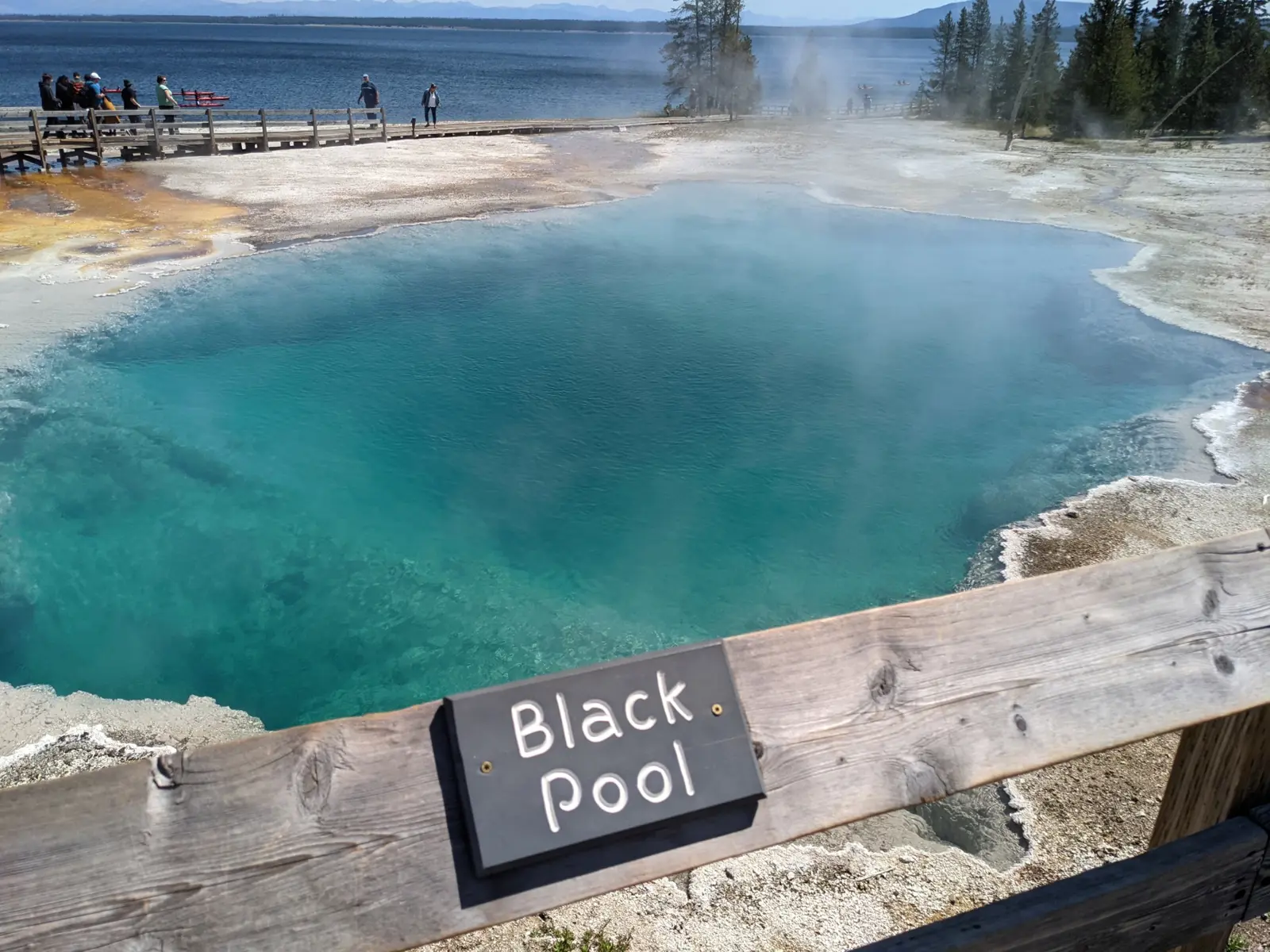 The teal water of Black Pool beside a wooden boardwalk with visitors, Yellowstone Lake stretching out behind