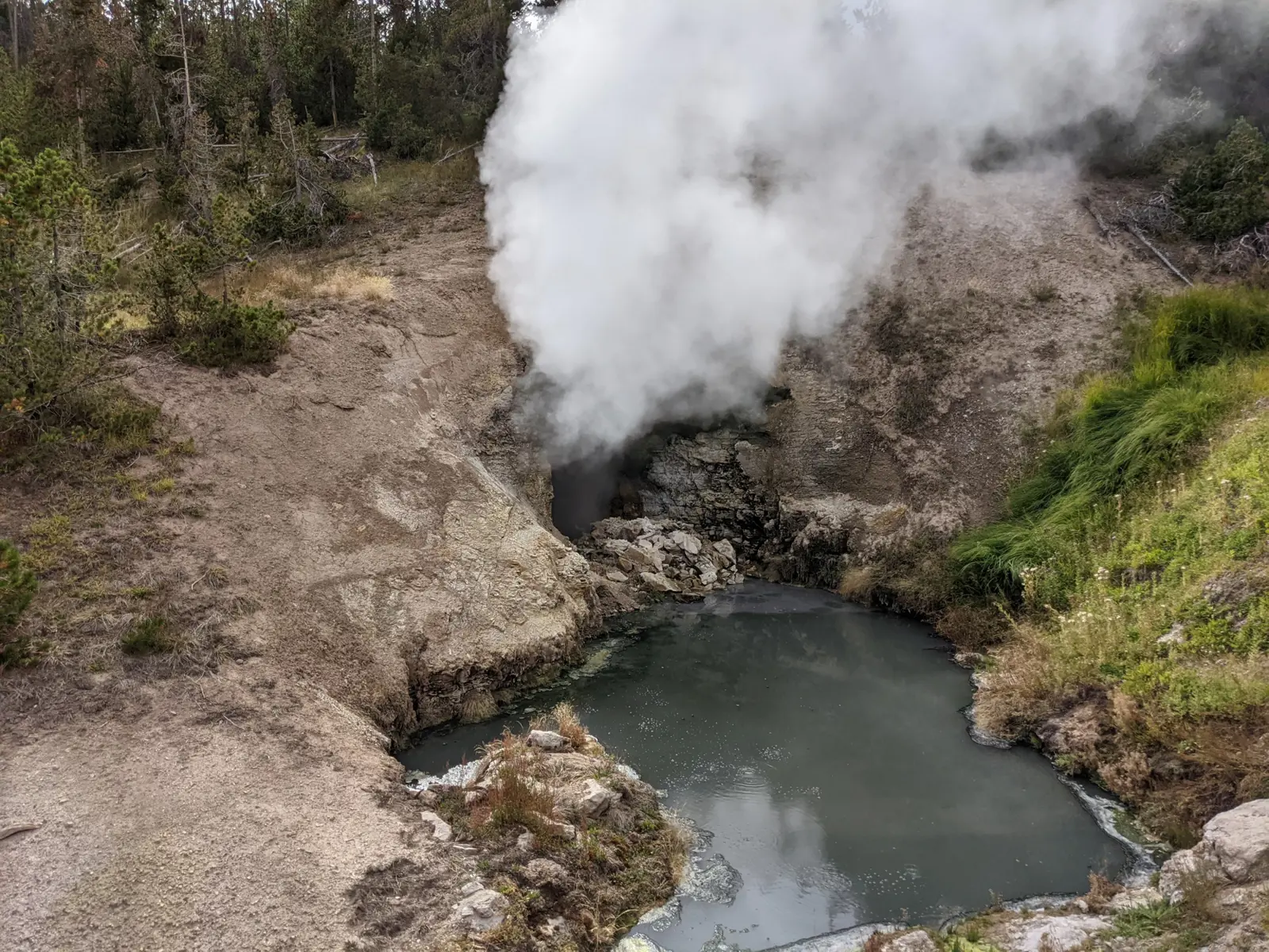 Steam pouring from a dark cave mouth above a grey pool at Dragon's Mouth Spring, surrounded by bare hillside