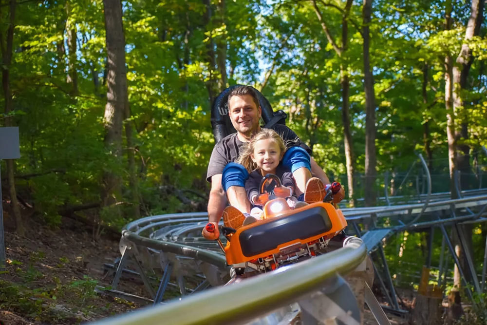 A smiling adult and young girl ride tandem in an orange mountain coaster cart on a steel rail winding through a sunlit forest