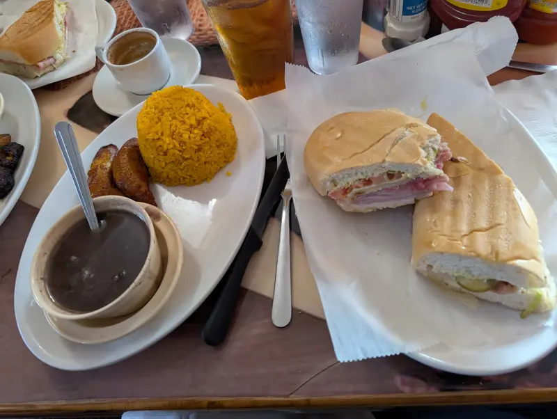 A Cuban sandwich cut in half on paper beside an oval plate of yellow rice, sweet plantains, and a bowl of black beans, with a small cup of Cuban coffee behind