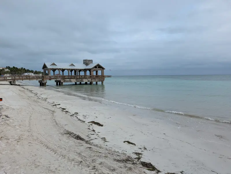 A covered wooden pier extending over calm pale-green water from a sandy beach under an overcast sky