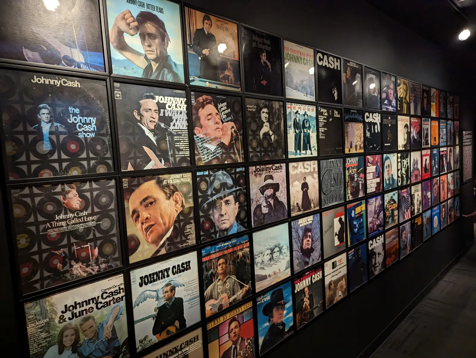 A long dark gallery wall lined edge to edge with framed vintage record album covers, receding toward a lit exit