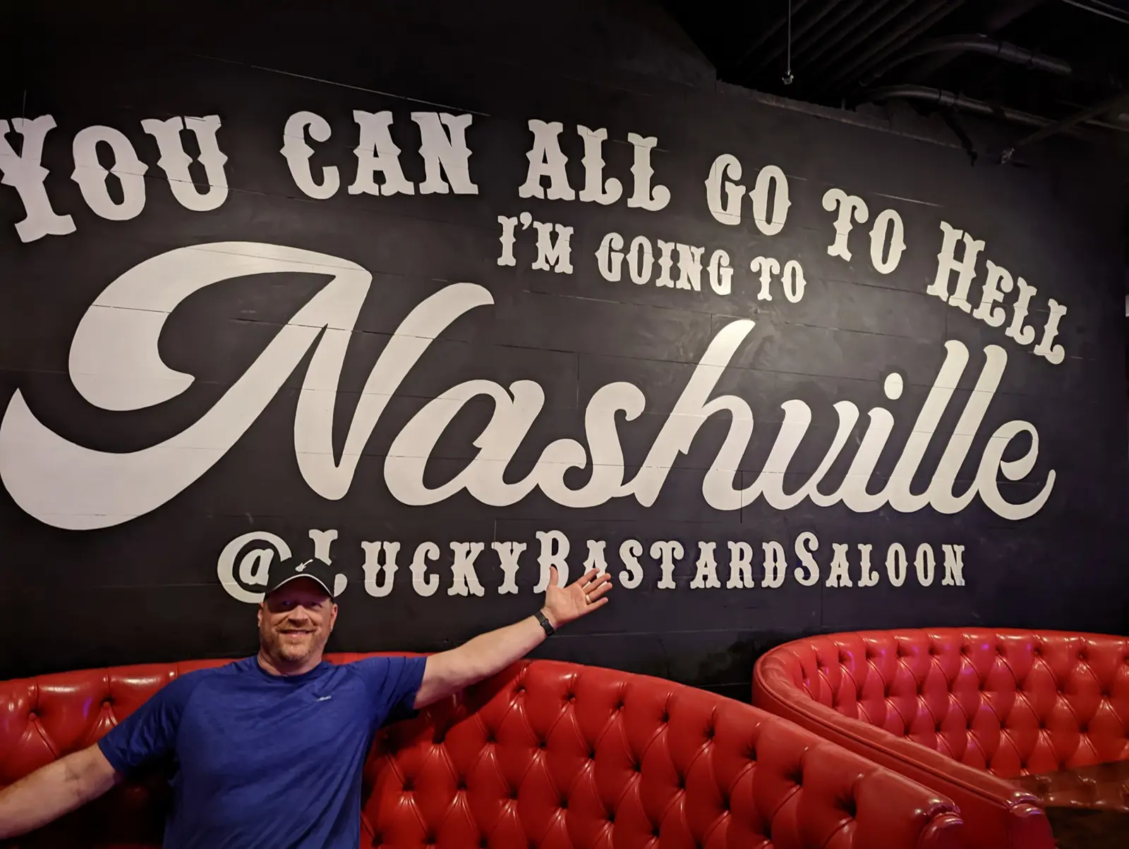 A man in a blue shirt and cap grinning with arms spread in front of a black wall painted with a giant Nashville script and red tufted leather booths