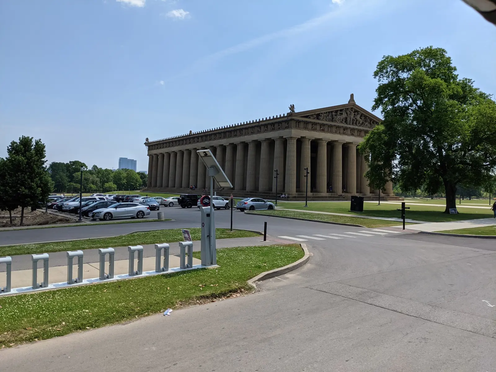 A full-size Doric temple with a carved pediment standing on a lawn beside a parking lot and bike-share dock under a hazy blue sky