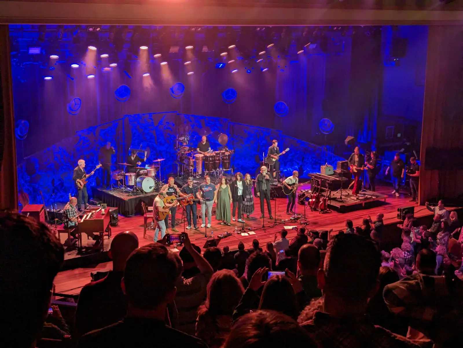 A crowded full-band encore on a wooden stage under blue and amber spotlights, seen from the balcony of a packed theater