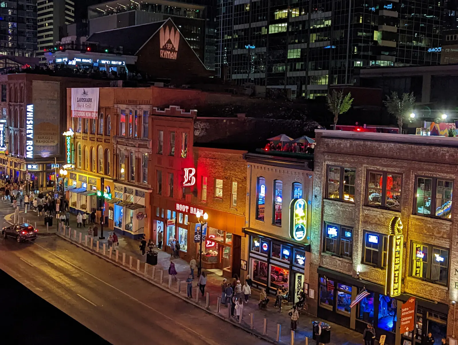 Lower Broadway at night seen from a rooftop, neon honky-tonk signs glowing along brick storefronts with the lit Ryman Auditorium gable behind