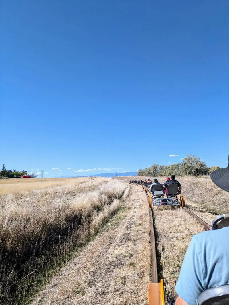 A convoy of four-seat rail bikes strung out along a single track through dry golden grassland under a deep blue sky, with a distant mountain range on the horizon