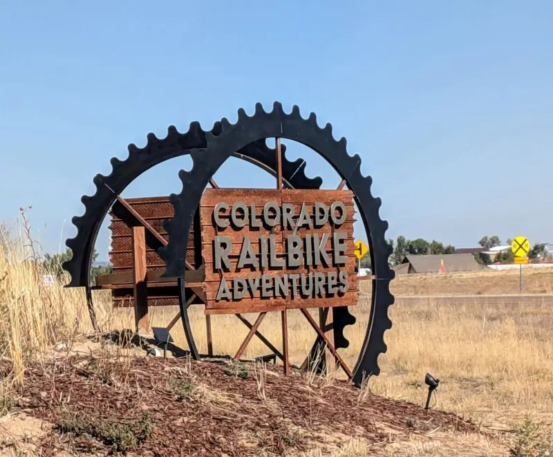 Roadside sign shaped like two black bicycle chainrings framing a rusted-metal panel reading Colorado Railbike Adventures, with a railroad crossing sign behind
