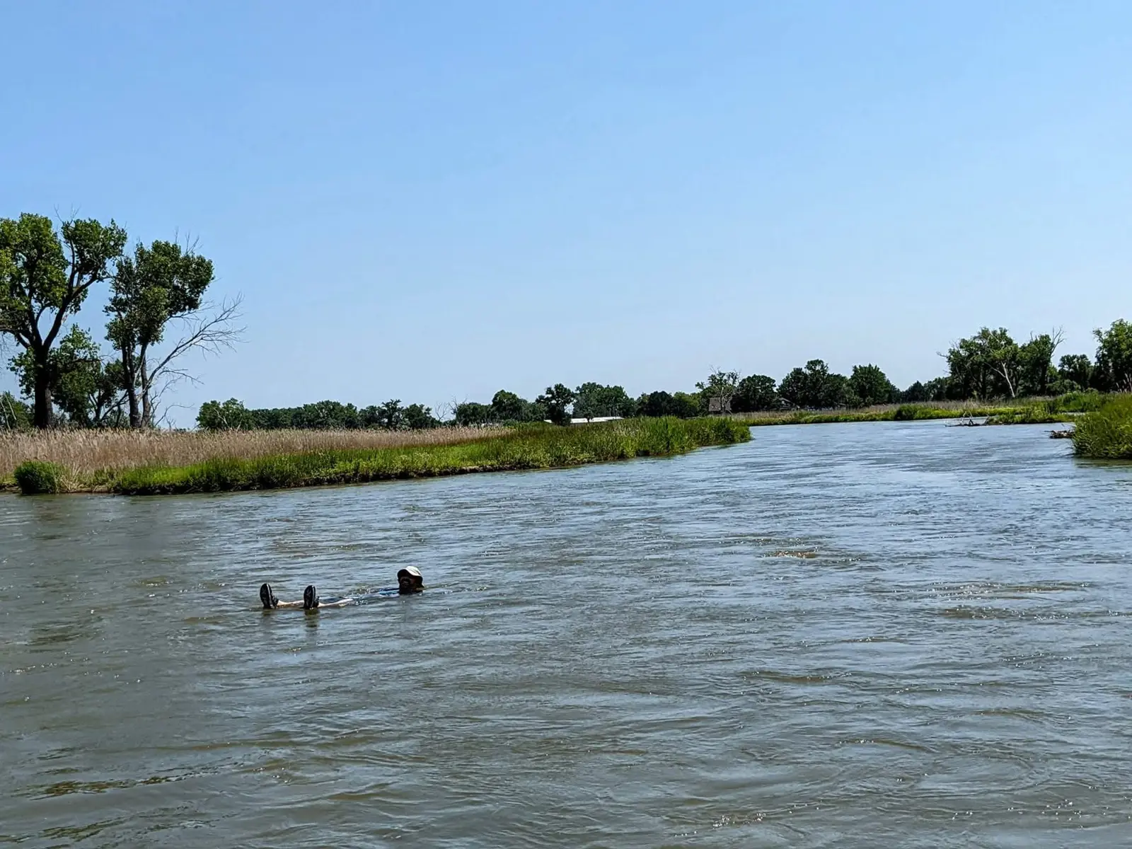 Wide, shallow Nebraska river lined with cottonwoods and tall grass under a clear blue sky, with two people wading in the water far downstream