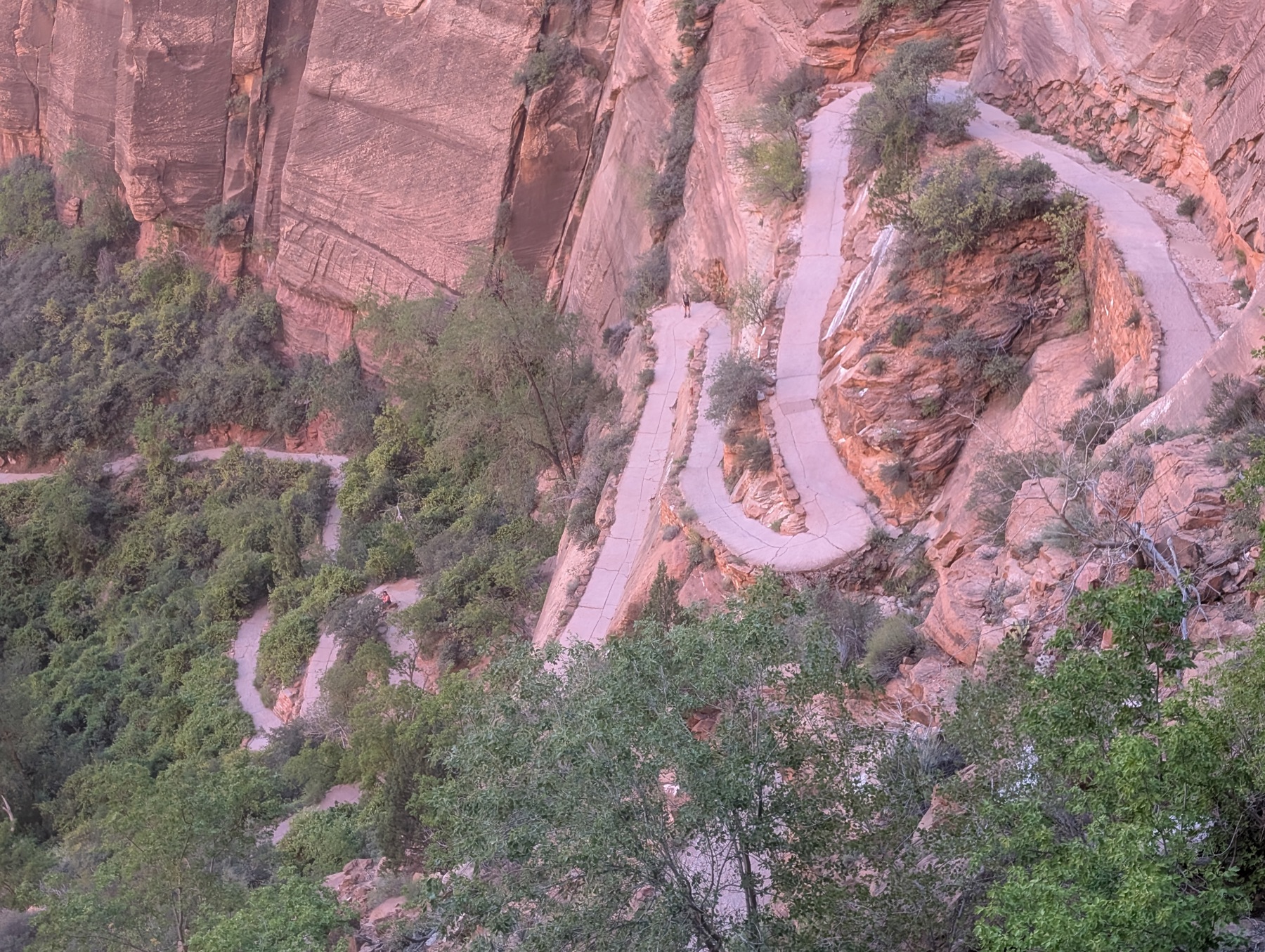 A paved trail zigzagging in tight stacked switchbacks up a sheer pink rock face, with a lone hiker on one of the turns