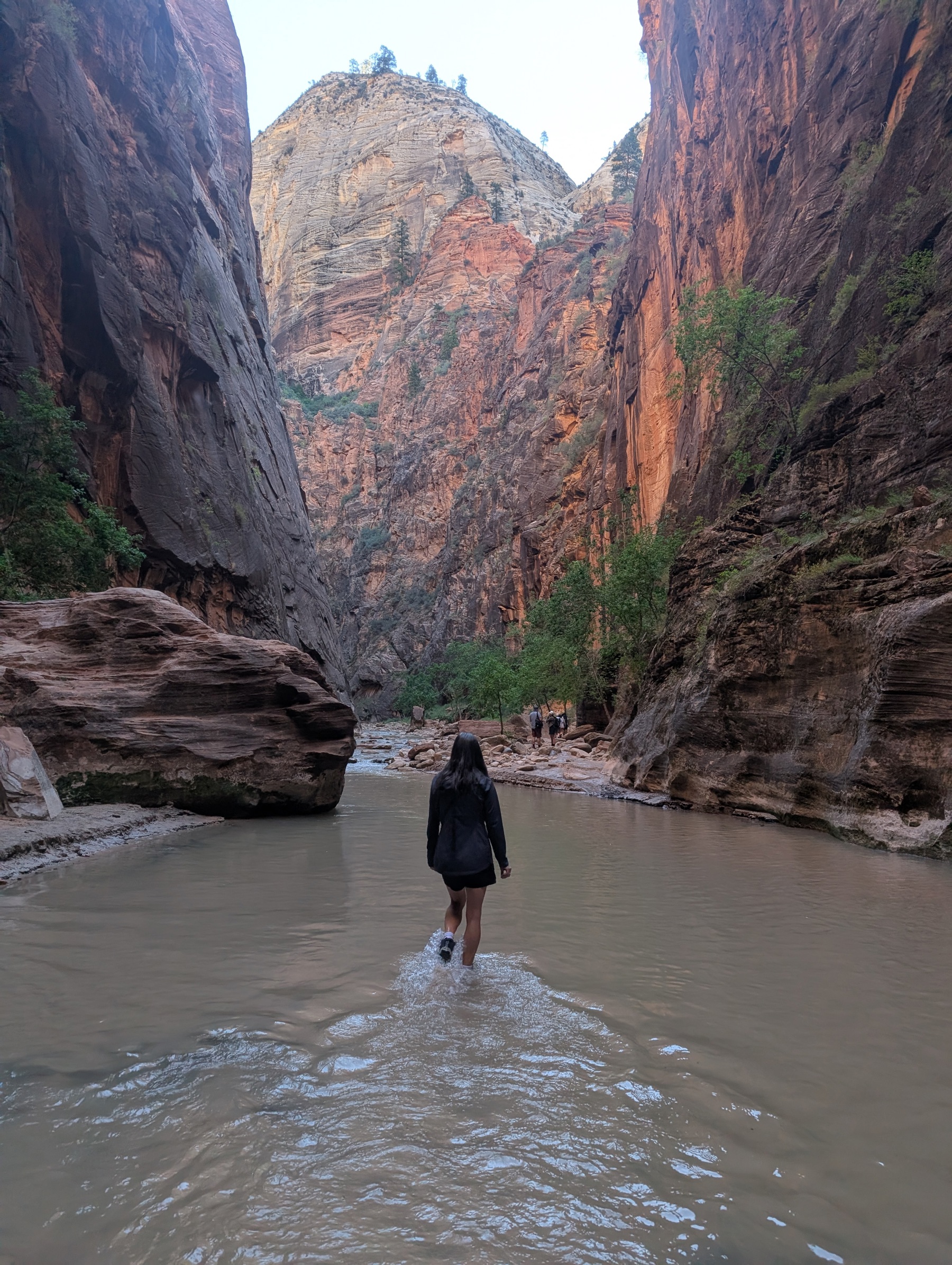 A hiker wading a shallow silty river between towering canyon walls, with a white-capped peak glowing in the distance