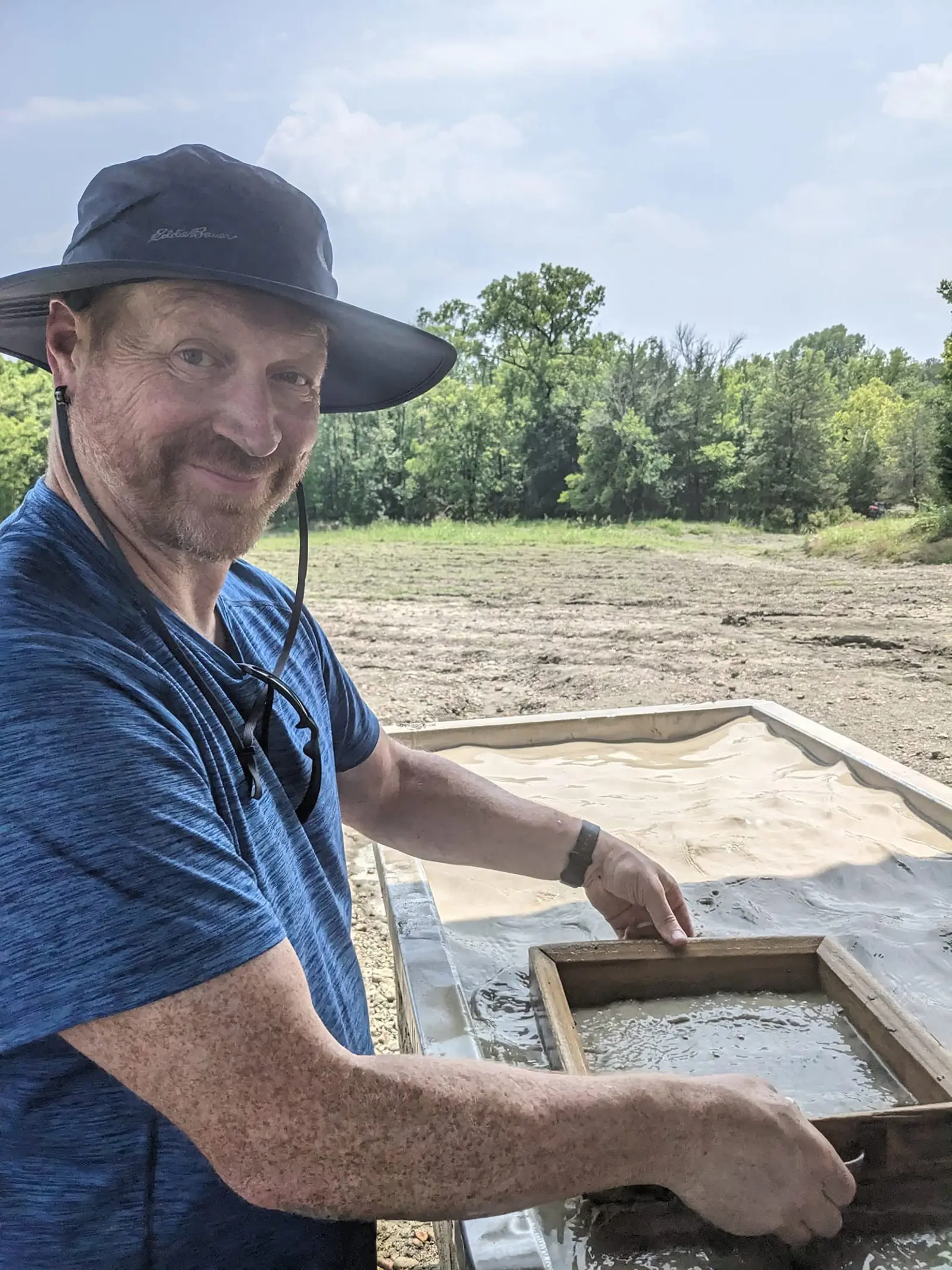 A smiling man in a wide-brimmed hat and blue T-shirt shaking a wooden screen of muddy gravel in a water-filled trough, with the bare, plowed search field and a tree line behind him