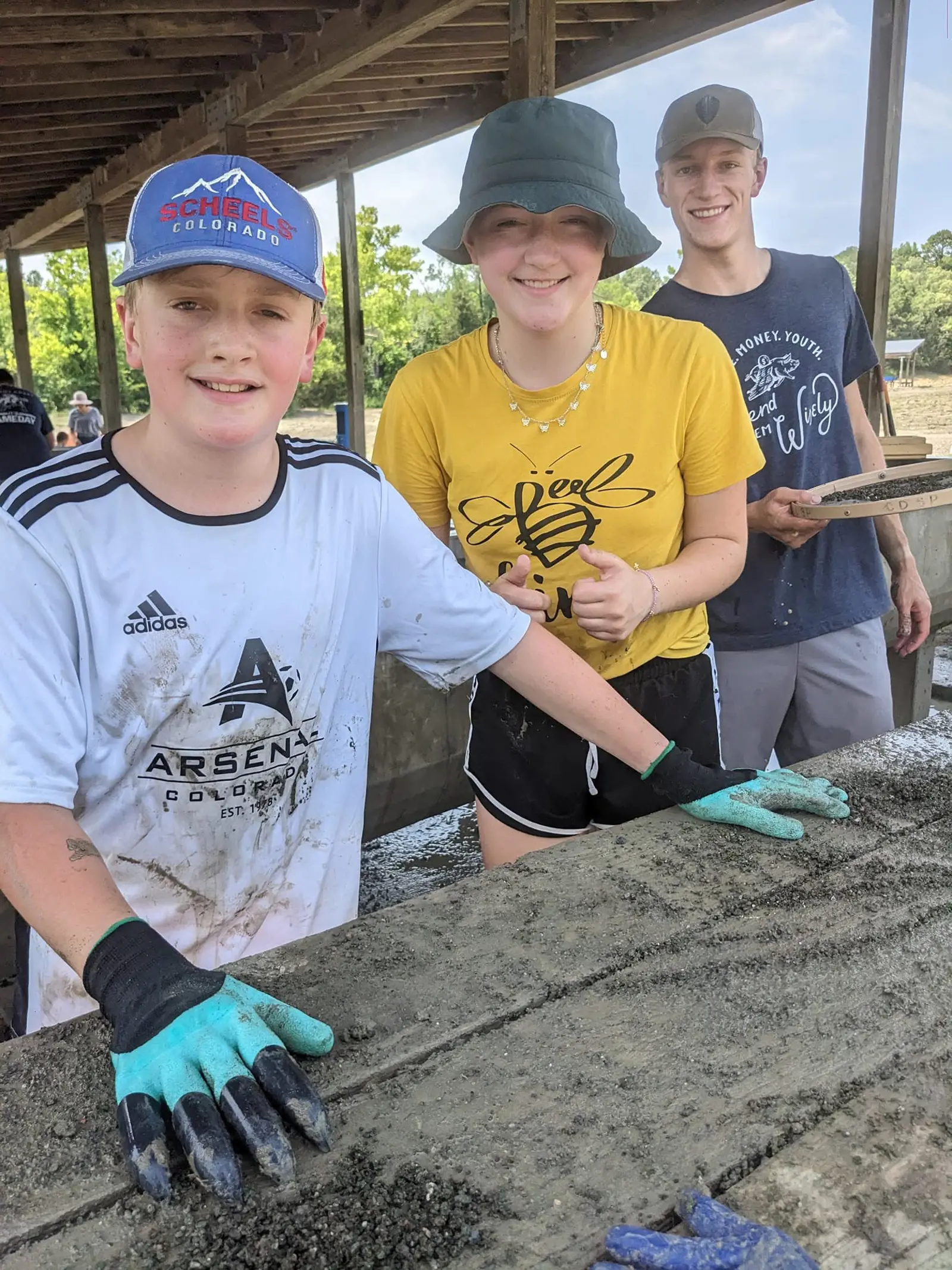 Three smiling young people in muddy T-shirts and garden gloves at a wooden sorting table spread with dark wet gravel, one holding a round sieve