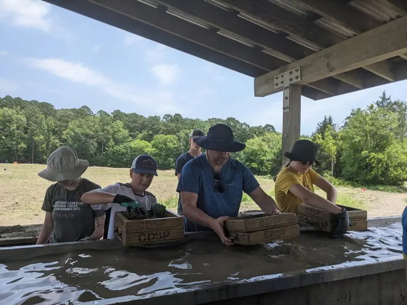 A family standing at a covered concrete water trough, washing gravel through wooden-framed screens, with the open dirt search field and a line of green trees behind them